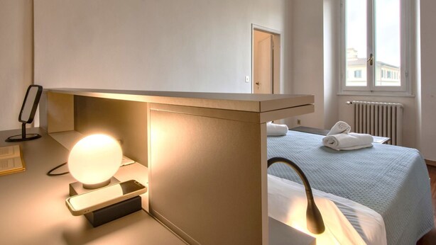 Hotelkamer met een bed, nachtkastje met lamp, spiegel en raam met uitzicht op gebouwen.