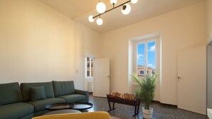 Een woonkamer met een bank, een salontafel, een zitbank met boeken, een bloempot en een groot raam met uitzicht op gebouwen.
