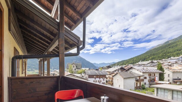 Un balcón con una mesa lista para dos personas, con vistas a un pueblo de edificios tradicionales y montañas al fondo.