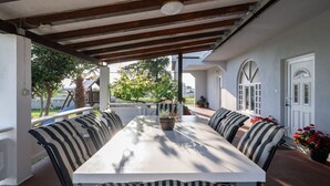 Une terrasse couverte avec une longue table et des bancs à rayures, entourée de murs blancs et d’un plafond en bois.