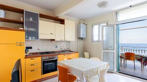 Een kleine keuken met gele kastjes, een eettafel met stoelen en uitzicht op zee door een glazen deur.