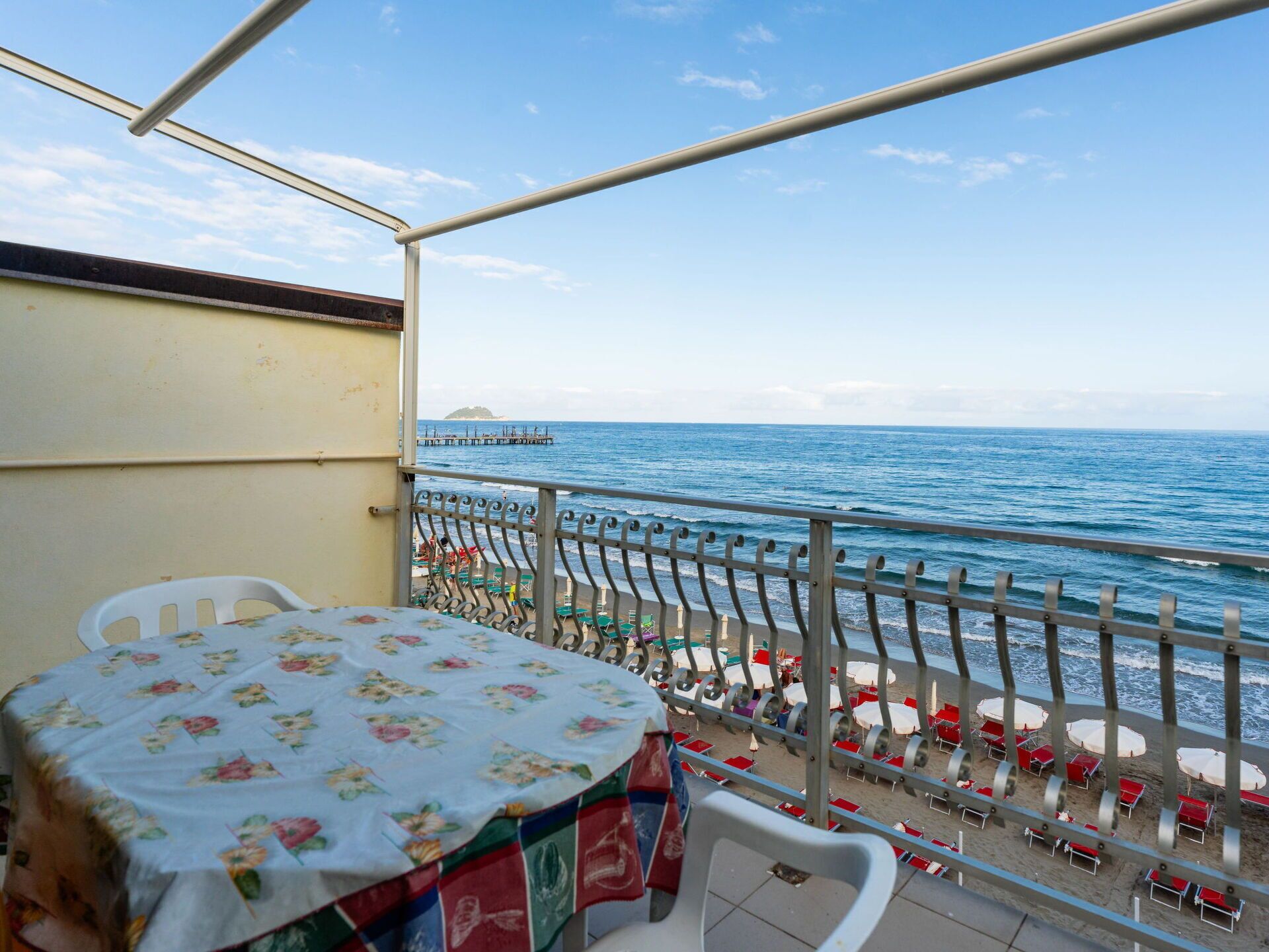 A balcony with a table and chairs, overlooking a beach with umbrellas and a pier.