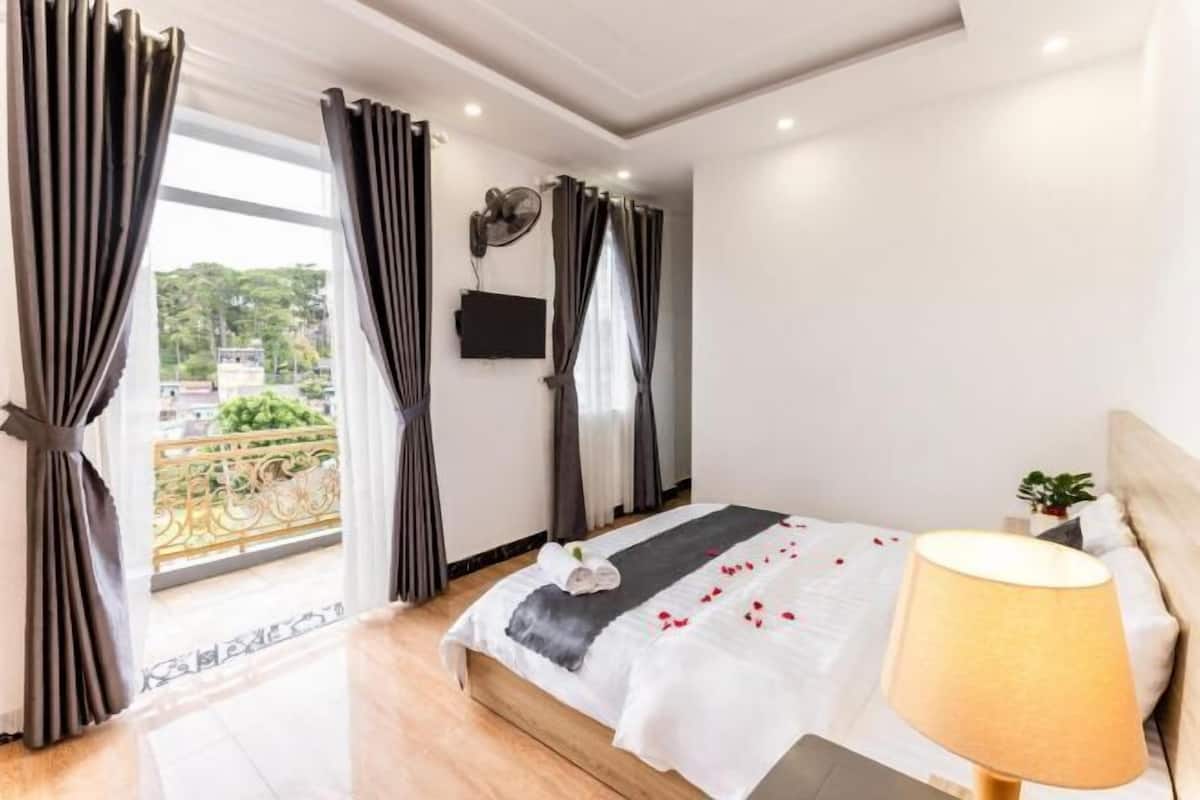 A bedroom with a bed, a bedside lamp, a TV on the wall, and a balcony with curtains.