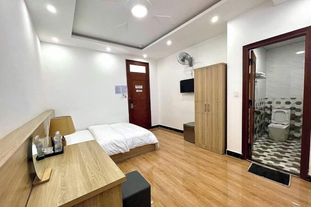 A hotel room with a bed, wooden bedside table, a wardrobe, a shower area, and a small desk with a chair.