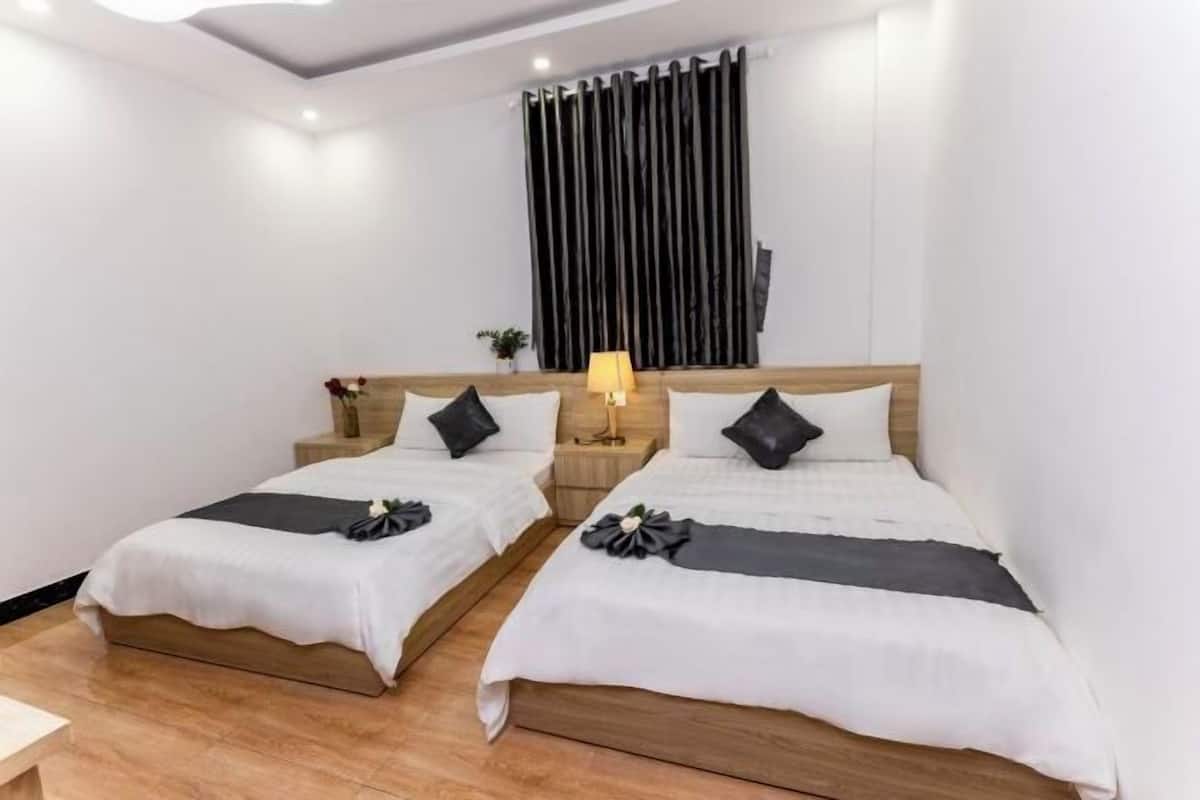 A hotel room with two single beds, wooden headboards, white bedding, a nightstand with a lamp, and a window with vertical blinds.
