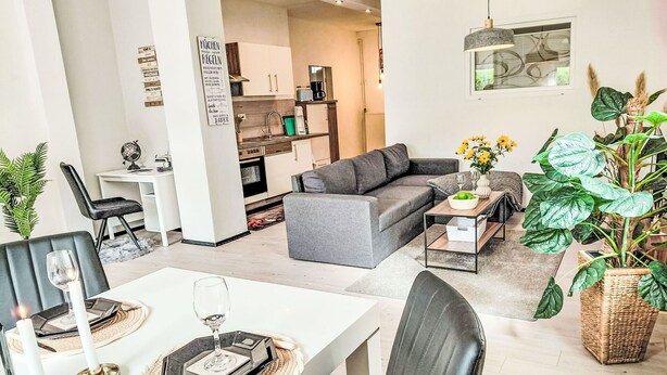 A modern living room with a grey sofa, a dining table set for two, and a kitchen in the background.