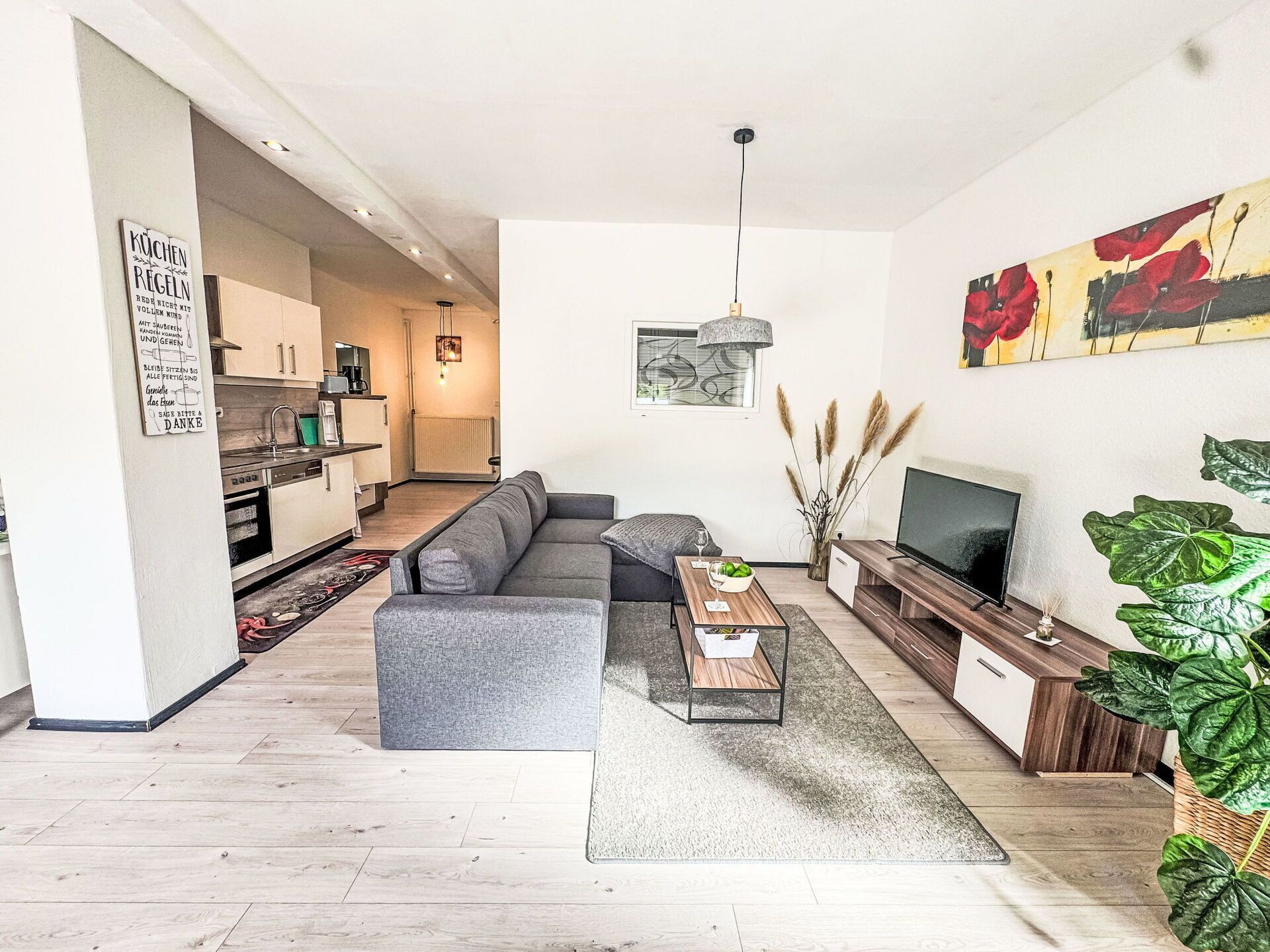 A modern living room with a grey sectional sofa, a coffee table, and a TV stand. There is a kitchen visible in the background with white cabinets and a hanging light fixture.
