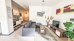A modern living room with a grey sectional sofa, a coffee table, and a TV stand. There is a kitchen visible in the background with white cabinets and a hanging light fixture.