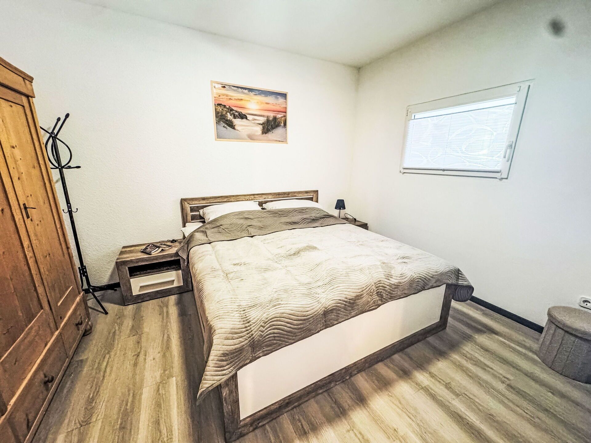 A bedroom with a wooden bed, a nightstand, a lamp, a wooden wardrobe, and a framed picture on the wall.