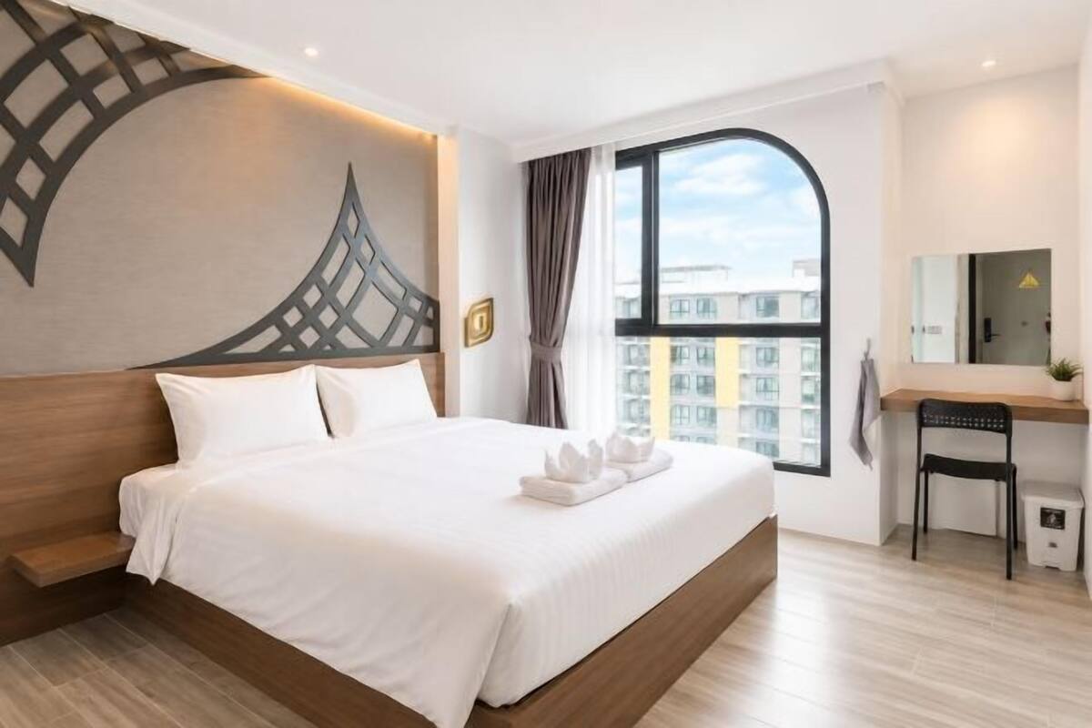 A hotel room with a large bed, a desk, a chair, a window with a view of buildings, and a decorative wall piece.