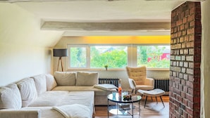 Una sala moderna con un sofá seccional, una mesa de centro redonda, un sillón y vistas a zonas verdes al exterior.