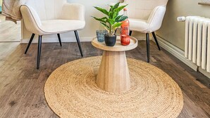 Una habitación con dos sillones color beige, una mesa de centro redonda de madera, un tapete tejido, una planta en maceta, un vaso y una botella de zumo de toronja rosa.