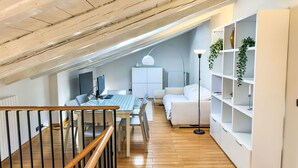 A modern loft bedroom with a wooden ceiling, a dining area with a glass table, a white sofa, and a bookshelf.