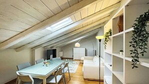A dining area with a table and chairs, a white sofa, a floor lamp, and a bookshelf with plants.