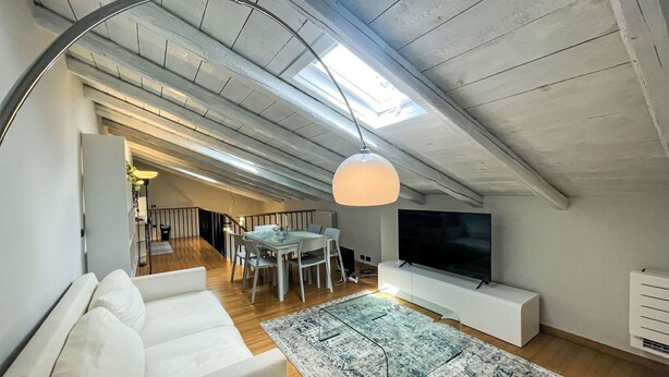 A modern loft apartment with a high ceiling, a dining area, a living room with a sofa and TV, and a large window providing natural light.