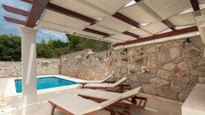 An outdoor pool area with stone walls, wooden lounge chairs, and a pergola structure.