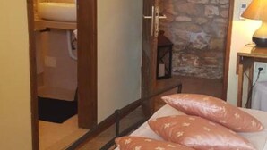 Un dormitorio con cama de madera, pared de piedra, una mesita con lámpara y un baño visible a través de una puerta abierta.