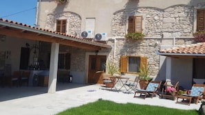 A two-story building with stone walls, wooden shutters, and a tiled roof. There are outdoor seating areas with chairs and tables, and a small garden area with potted plants.
