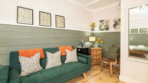 Un sofá verde con cojines estampados, una mesita de madera, una lámpara y un espejo en una habitación con paneles de madera y obras de arte en las paredes.