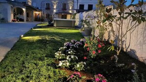 Un patio con césped cuidado y macizos de flores, un edificio de piedra con balcón, y una jardinera grande con un árbol.