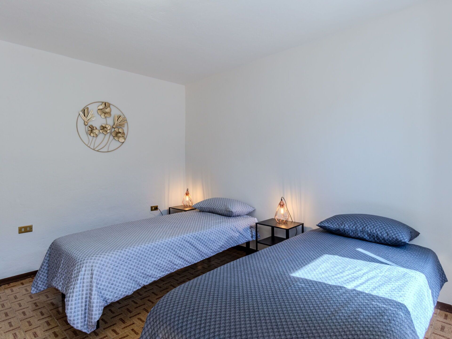 A room with two single beds, each with a patterned bedspread and a matching pillow. There are two bedside tables with lamps, and a decorative wall piece above the beds.