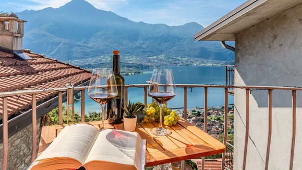 Una mesa de madera con dos libros abiertos, dos copas de vino, una botella de vino y un racimo de uvas en un balcón con vistas a un lago y montañas.