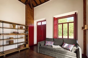 A room with a wooden floor, a grey sofa with patterned cushions, a red door, and a window with a view of greenery. - Studio dos Cantoneiros (Salir)