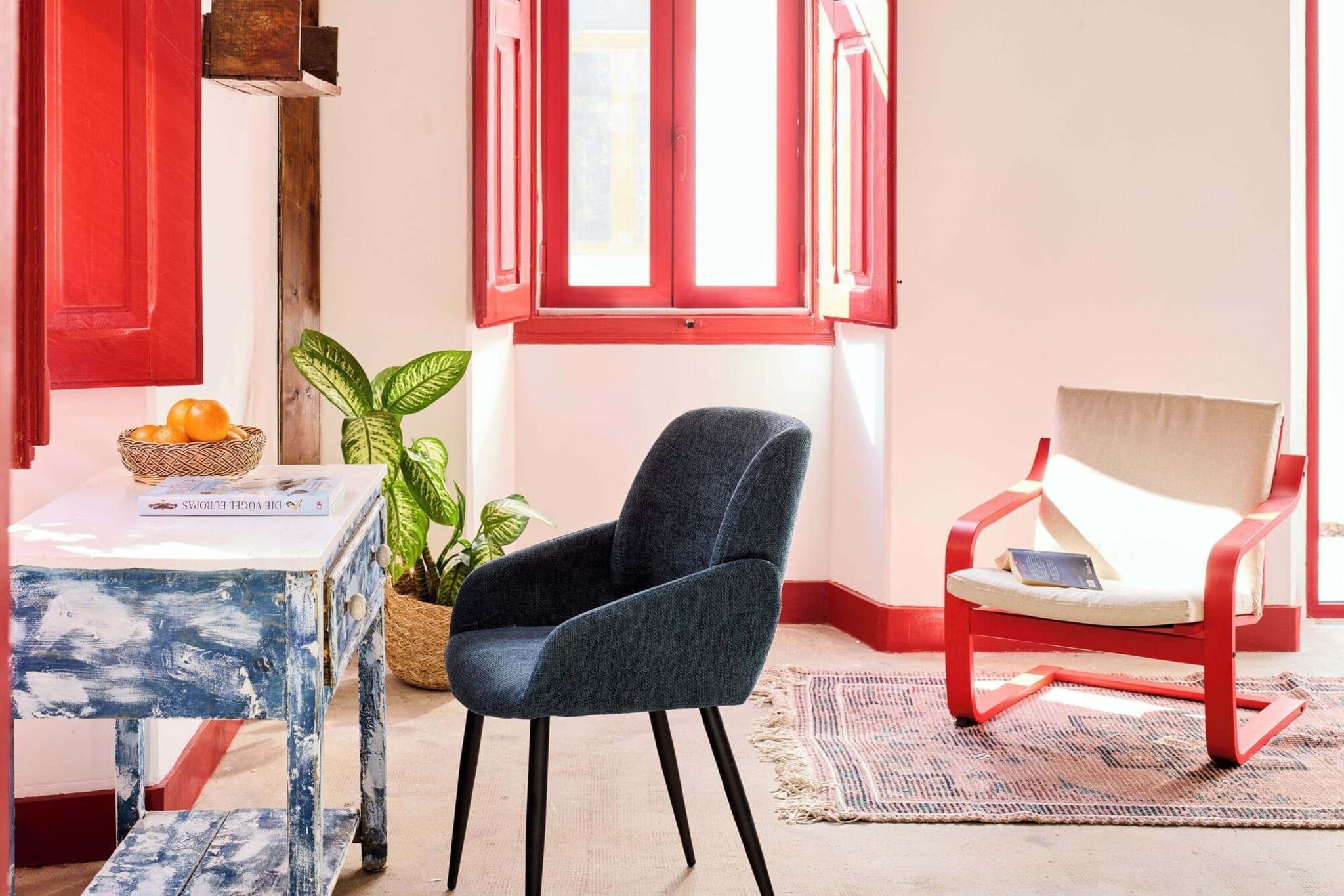 Een kamer met een blauwe stoel, een rode stoel, een tafel met een mand sinaasappels en een kamerplant.