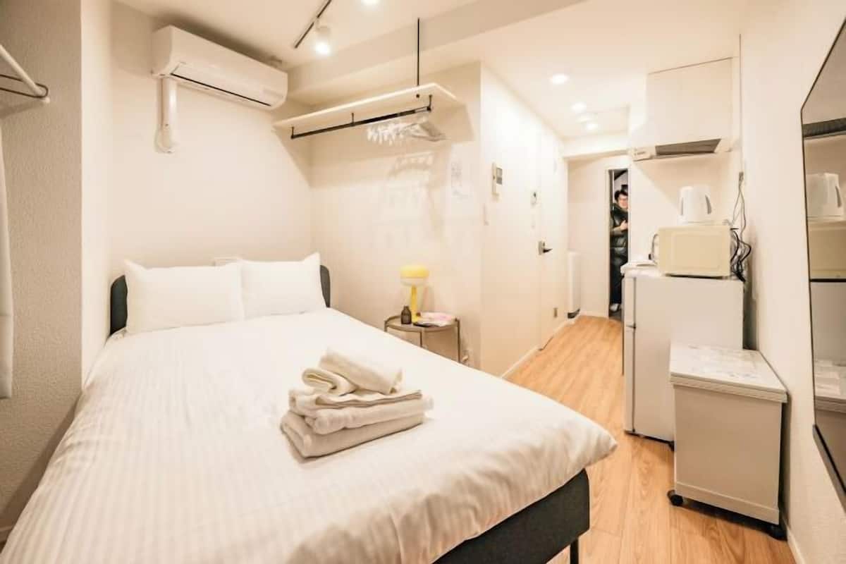 A hotel room with a bed, a bedside table with a lamp, a small cabinet, and a kitchenette area.