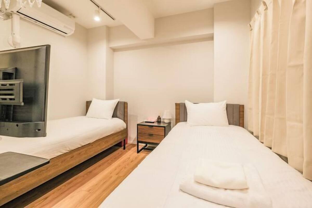 A modern hotel room with two single beds, a wooden floor, a small bedside table, and a wall-mounted air conditioning unit.