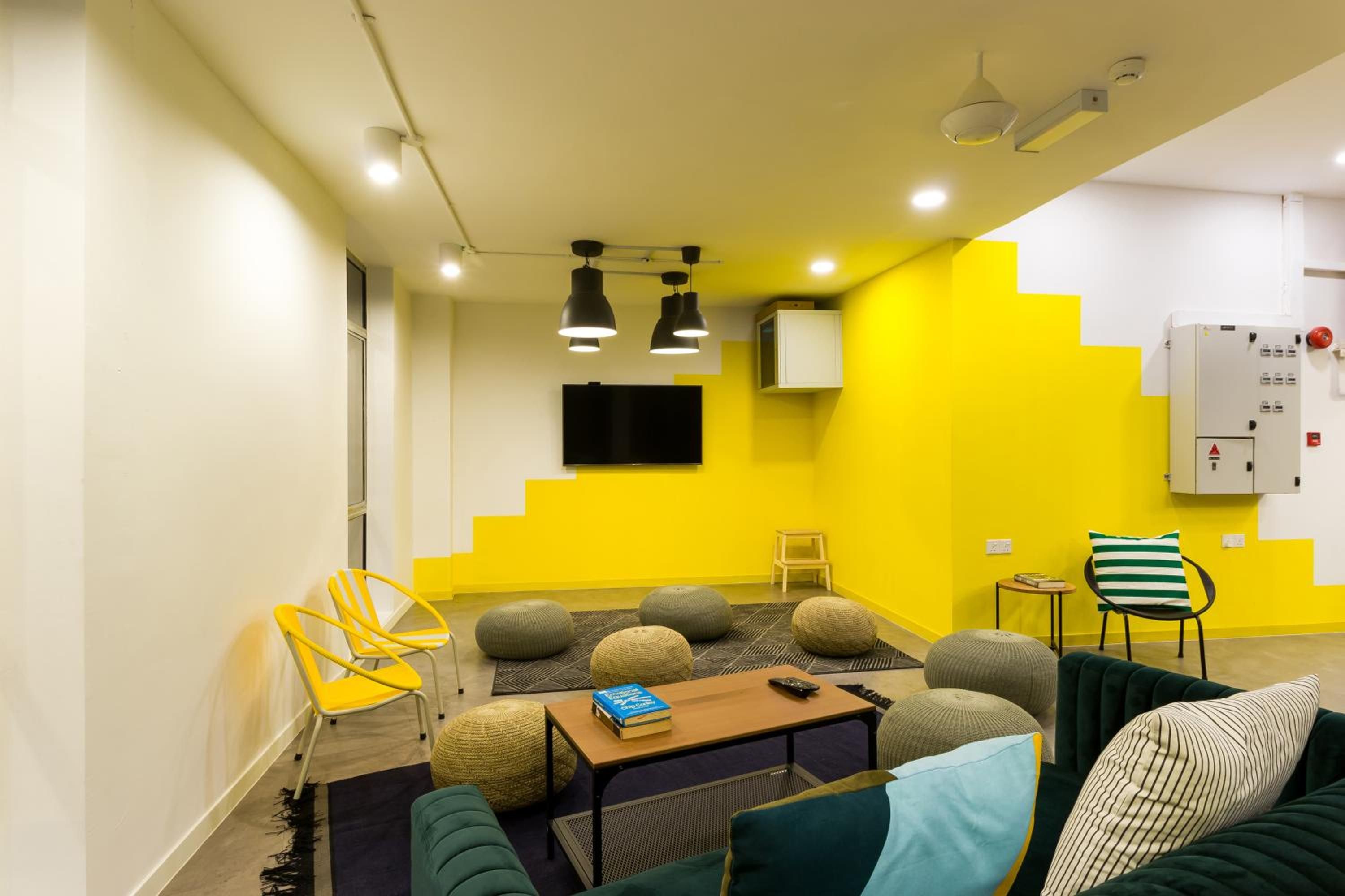 Una sala de estar moderna con una pared de acento amarilla, un televisor de pantalla plana montado en la pared, una mesa de centro de madera con libros y un control remoto, y un sofá verde con cojines a rayas.