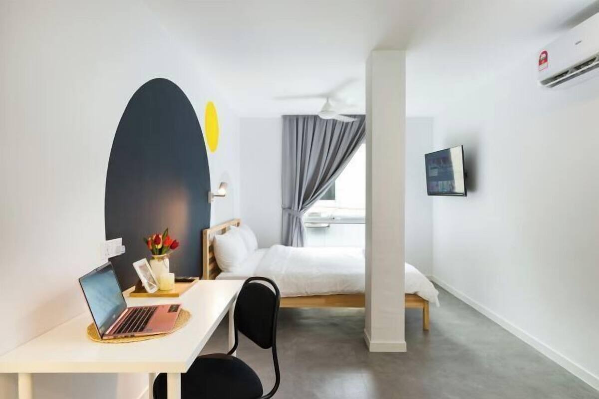 Ein Hotelzimmer mit einem Bett, einem Schreibtisch mit Laptop, einem Sessel, einem Fernseher und einem Fenster mit Vorhängen.