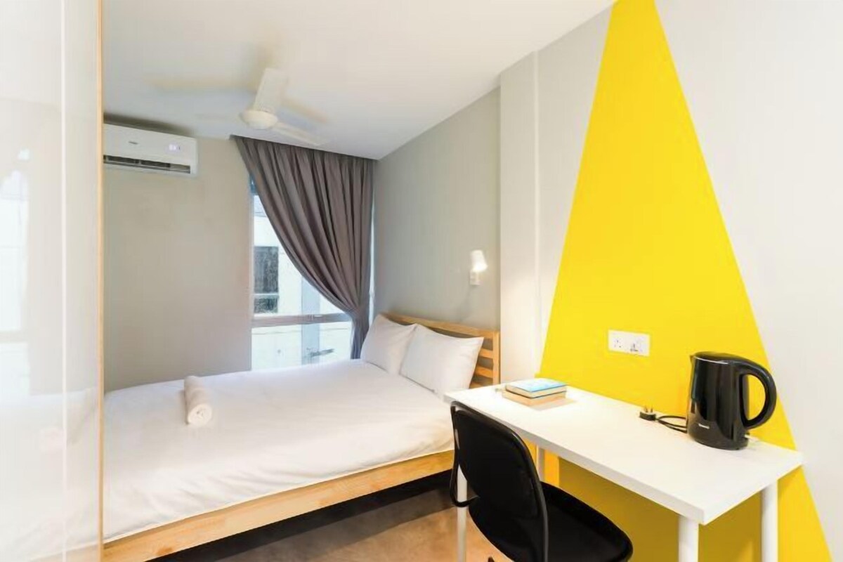 A hotel room with a bed, a desk, a chair, a kettle, and a window with curtains.