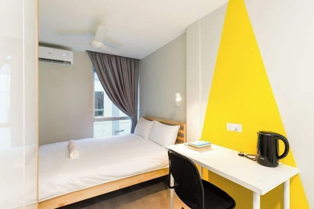 A hotel room with a bed, a desk, a chair, a kettle, and a window with curtains.
