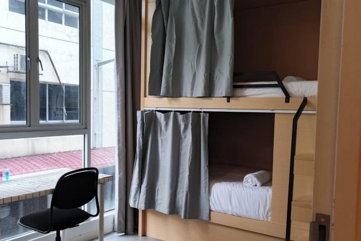 A dormitory room with bunk beds, a window with curtains, and a chair by the window.