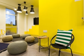 A room with a yellow wall, a black chair with a striped pillow, and several round ottomans. - Upstairs Downstairs Coliving Kampung Pandan (Kuala Lumpur)