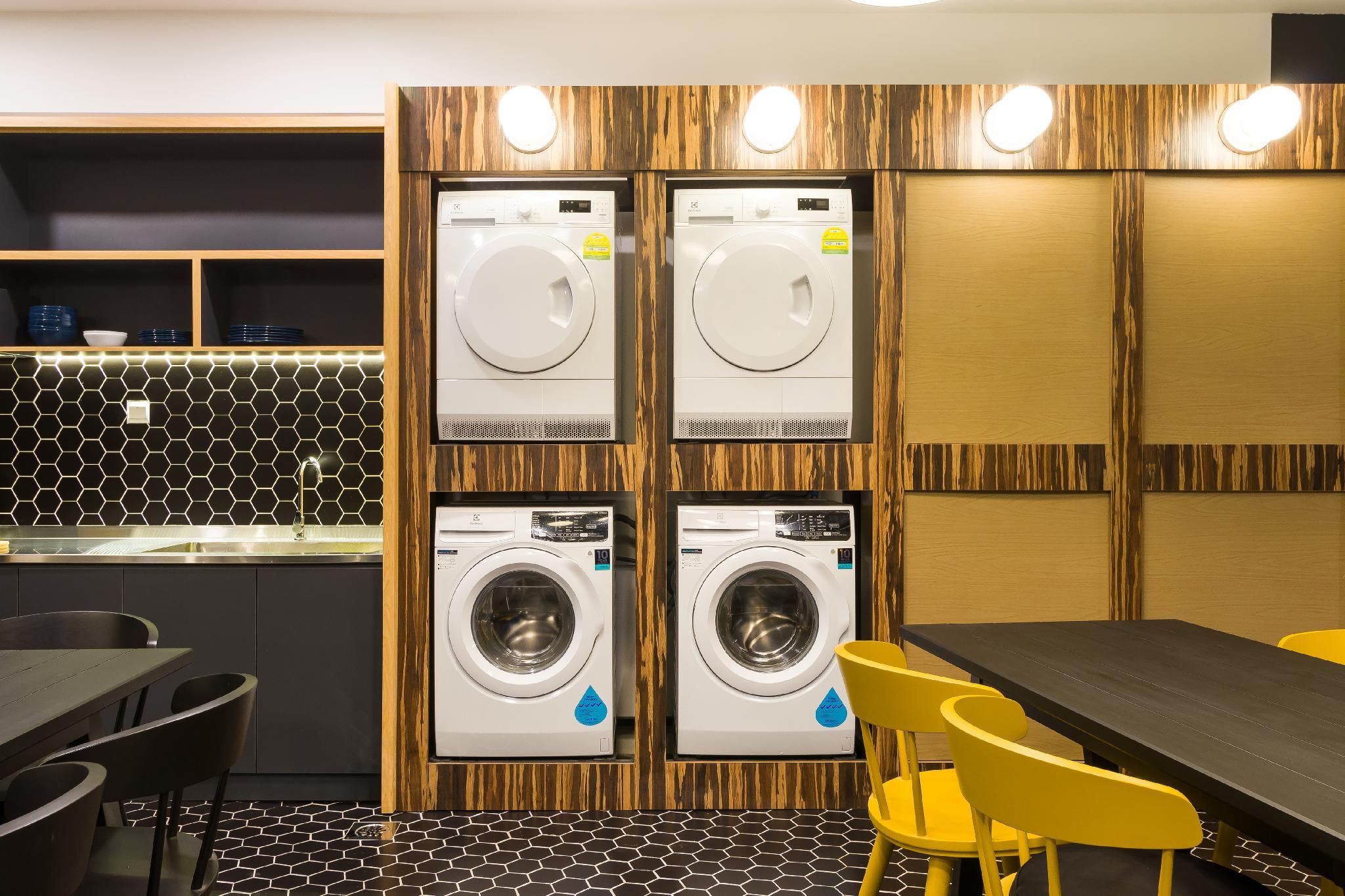 A modern laundry room with four washing machines, a sink, and a dining area with yellow chairs.