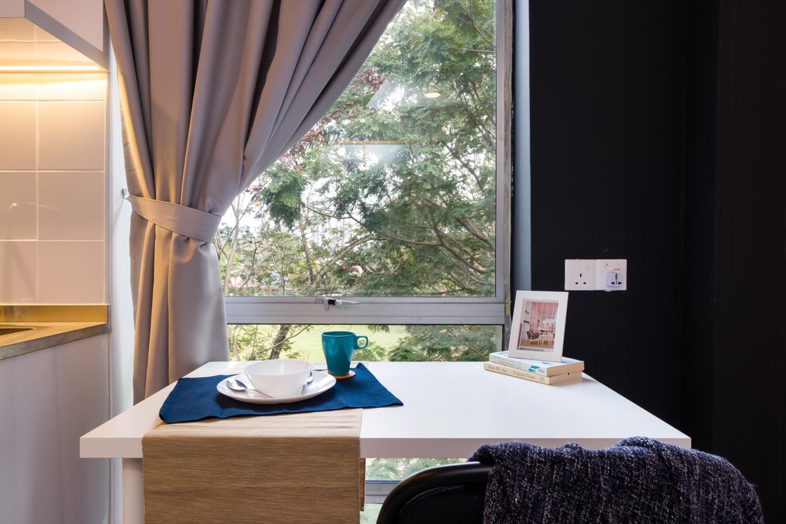 Una habitación con ventana, una mesa con un plato, una taza y un cuadro enmarcado.