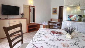A dining area with a table set for two, a television mounted on the wall, and a kitchen area with cabinets and a stove. - Apartments Butković - One Bedroom Apartment (A2) (Novi Vinodolski)