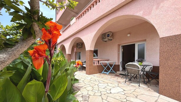 An outdoor patio area with stone flooring, a table and chairs, and a building with pink walls and arches. - Apartments Butković - One Bedroom Apartment (A2) (Novi Vinodolski)