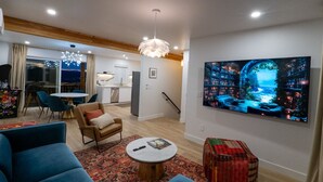 Una sala de estar moderna con un televisor de pantalla plana grande, un área de descanso acogedor con un sofá y sillones, una mesa de comedor y una cocina al fondo.