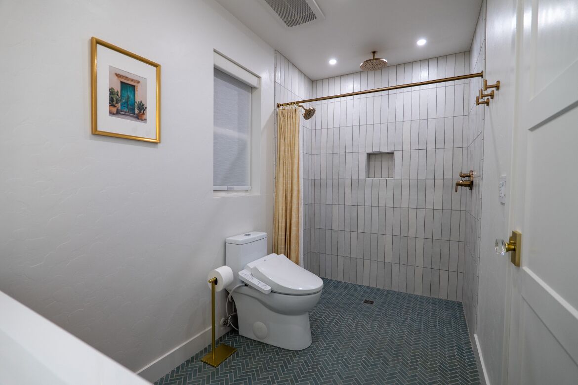 A bathroom with a toilet, a shower with a curtain, a framed picture on the wall, and a window.