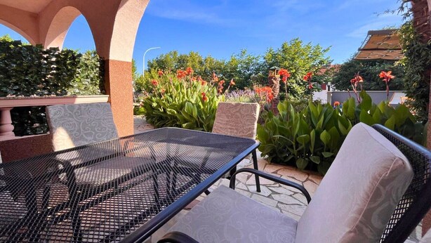 An outdoor patio with a metal table and cushioned chairs, surrounded by lush greenery and flowering plants.