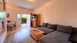 A living room with a sofa, a coffee table, and a dining area with a table and chairs. There is a bookshelf and a door leading outside.