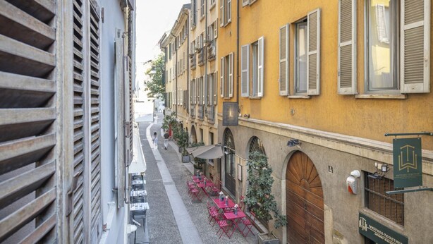 A narrow street with yellow buildings, outdoor seating, and a sign for 'Watches Together'. - Easylife - Milano - Carpoforo 5 - Brera (Milano)