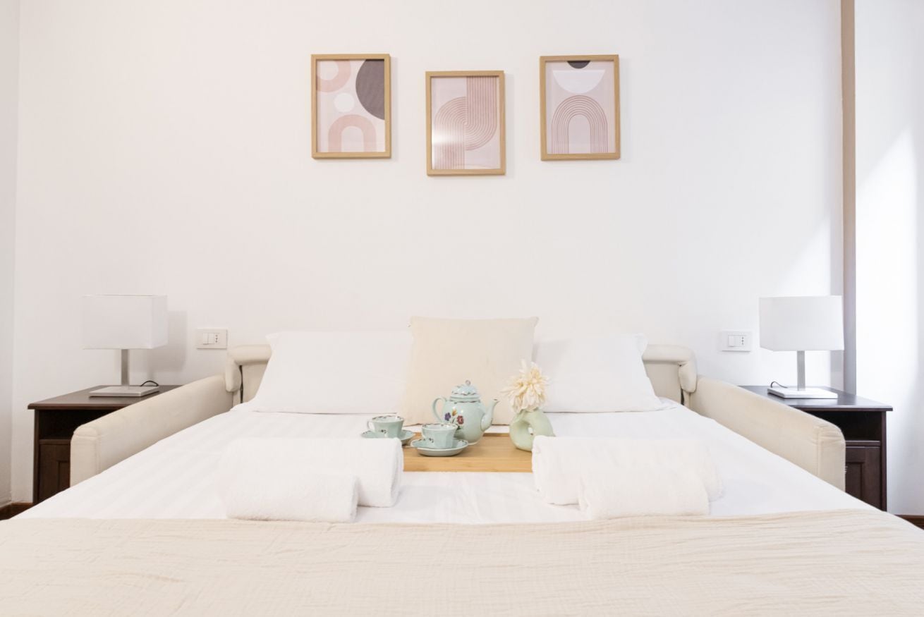 Habitación de hotel con una cama grande, dos mesitas de noche, una mesilla auxiliar y una pequeña mesa con juego de té. Hay tres cuadros enmarcados en la pared sobre la cama.