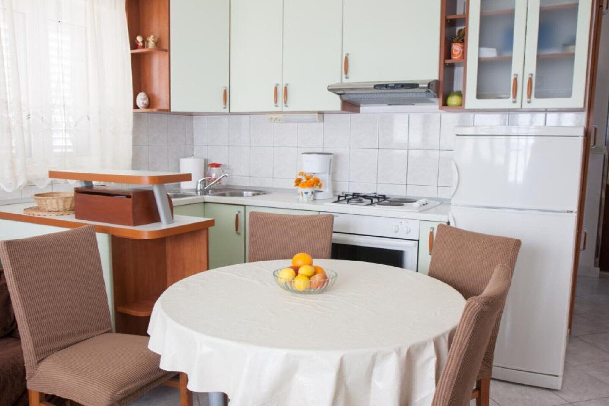 Cocina compacta con zona de comedor, que incluye una mesa redonda con un tazón de fruta, sillas de madera y armarios blancos.