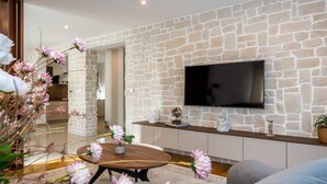 A modern living room with a stone wall, a flat-screen TV mounted above a wooden console, and a round coffee table with a vase of flowers. - Villa Branka - Villa Tomić Drniš - Siverić (Drnis)