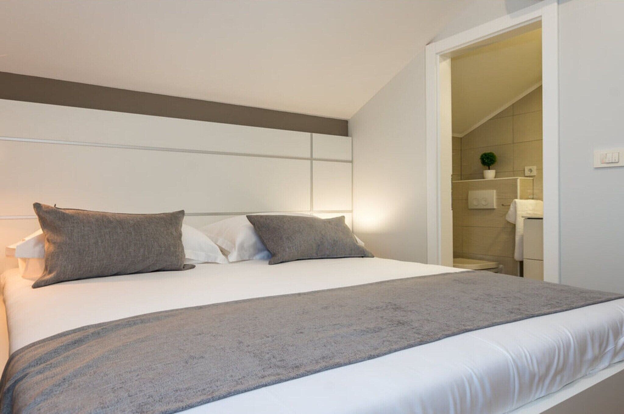 Un lit bien fait, avec une couverture grise et des oreillers, une tête de lit blanche, et une salle de bain visible, équipée d’un porte-serviettes et d’une plante en pot.