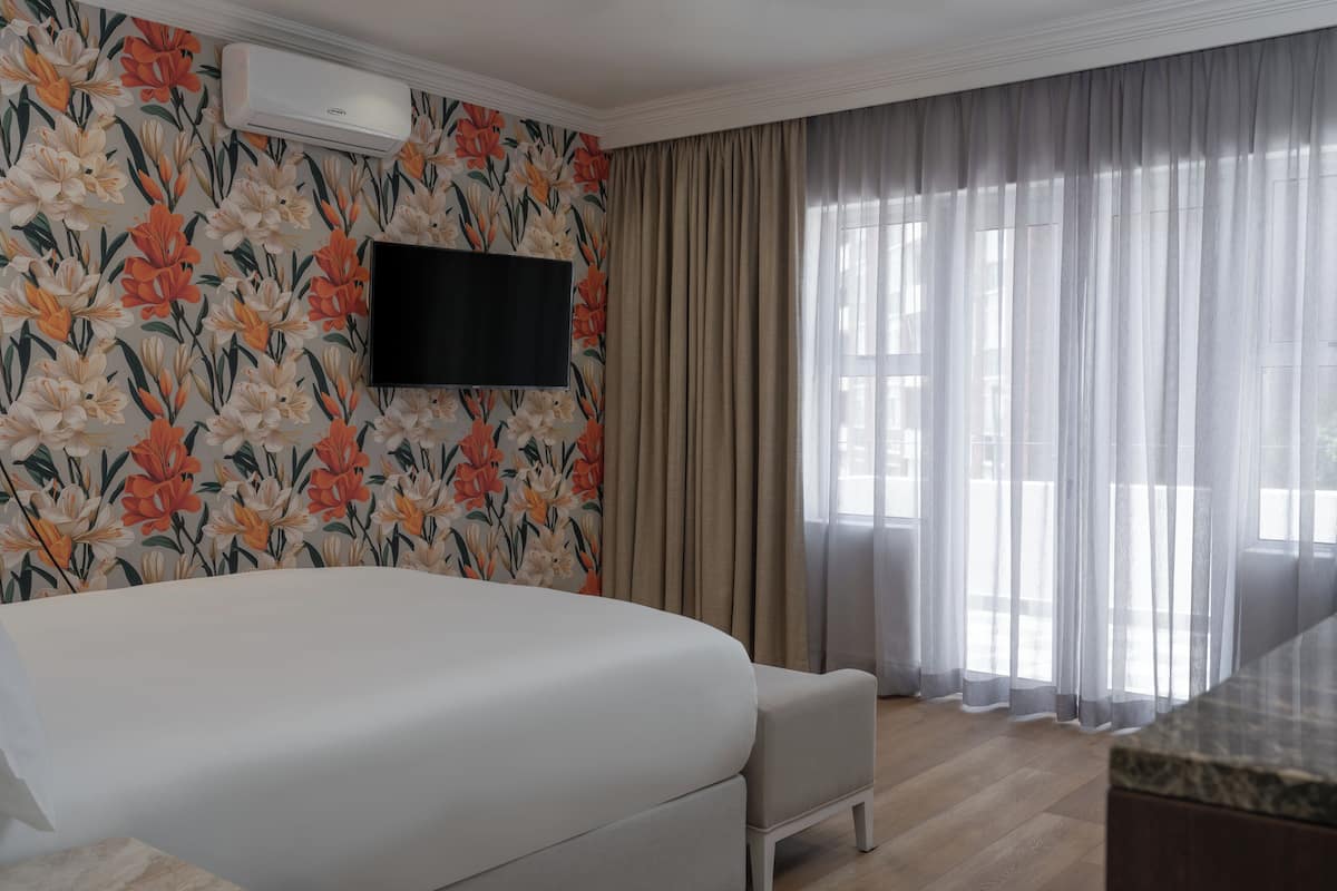 A hotel room with a large bed, a flat-screen TV mounted on the wall, and a large window with sheer curtains.
